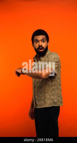 vue verticale d'un jeune homme afro-américain choqué avec montre-bracelet regardant la caméra sur orange, image de stock Banque D'Images