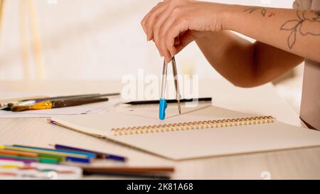 Vue rognée de la femme avec tatouage tenant la boussole de dessin près de l'esquisse à la maison, image de stock Banque D'Images