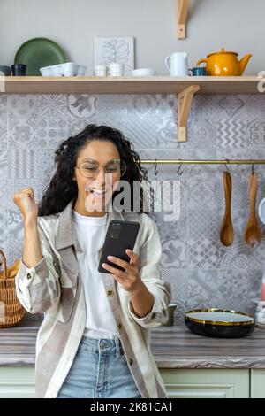 Vertical shot, une femme latino-américaine à la maison dans la cuisine a eu de bonnes nouvelles en ligne, une femme avec des cheveux et des lunettes mauries célébrant la victoire succès en tenant un smartphone. Banque D'Images