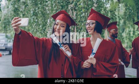 Les jolies filles qui terminent leurs études prennent le selfie avec des parchemins de diplôme à l'aide d'un smartphone, les jeunes femmes posent et sourient, leurs camarades célèbrent en arrière-plan. Banque D'Images