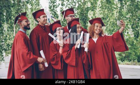 Les jeunes diplômés enthousiastes prennent le selfie avec leur smartphone, les jeunes font des diplômes, posent, sourient et rient. Éducation, jeunesse et réussite. Banque D'Images