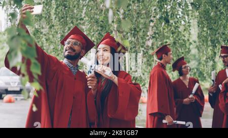 Deux étudiants prennent le selfie avec des diplômes de diplôme portant des mortarboards et des robes, Guy tient le smartphone et prend la photo, la fille pose. Banque D'Images