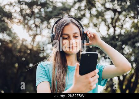 Une adolescente porte un t-shirt turquoise, utilise un casque et établit une liste de lecture musicale sur son smartphone dans le parc Banque D'Images