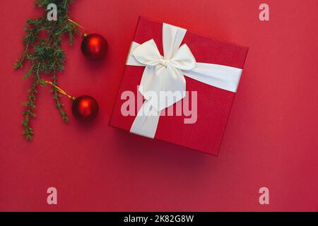 Boîte cadeau avec noeud en ruban et décorations de Noël sur branche d'arbre sur fond rouge. Jouets ronds. Style minimaliste. Copier l'espace. Préparation de cadeaux pour les vacances. Vierge pour la publicité Banque D'Images