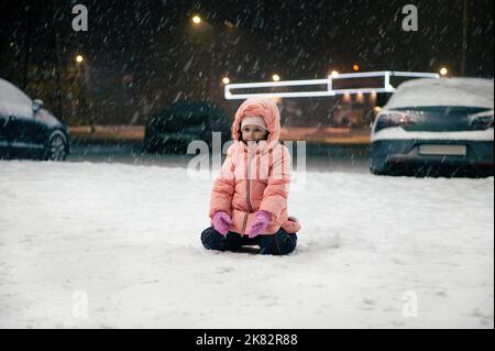 Un enfant heureux, une petite fille dans une combinaison chaude d'hiver s'assoit dans une rue enneigée, appréciant la première neige pendant une promenade en soirée Banque D'Images