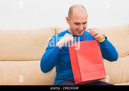 Un homme cachiste vêtu d'un T-shirt bleu à manches longues tient et ouvre un sac en papier rouge tout en regardant avec sourire à l'intérieur assis sur un Banque D'Images