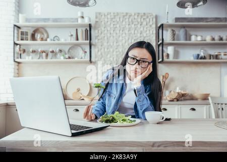 Insatisfait et fatigué de la diète jeune femme asiatique. Il est assis à la table dans la cuisine et regarde un séminaire en ligne sur un ordinateur portable. Il tient une salade fraîche, regarde dans l'appareil photo. Banque D'Images