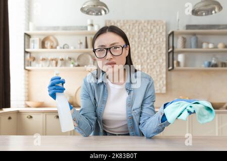 Jeune fatigué belle femme asiatique de nettoyage à la maison. Essuyer la table avec un chiffon dans des gants en caoutchouc et une bouteille de spray. Il regarde l'appareil photo, lui fait passer les mains. Banque D'Images