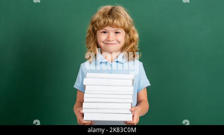 Enfant en classe. Un enfant heureux contre un tableau noir vert. Concept d'éducation et de créativité. Pile de livres pour enfants avec plaque de mortier Banque D'Images