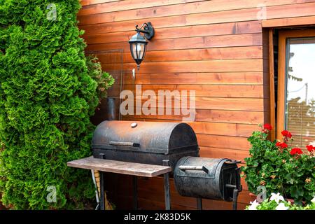 barbecue rouillé noir de fer près de la maison en bois avec lanterne rétro sur le mur fait de planches, cottage avec des buissons de thuja et des fleurs de pélargonium rouge, lieu à Banque D'Images