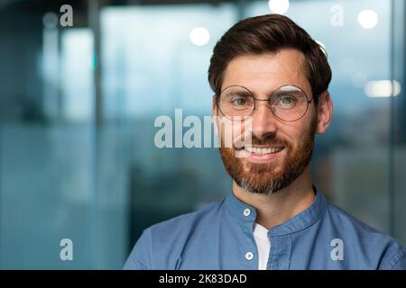 Homme d'affaires joyeux et réussi avec une barbe et des lunettes regardant l'appareil photo et souriant, portrait en gros plan de l'homme dans un bureau moderne. Banque D'Images