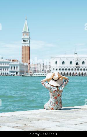 Vue de derrière une femme d'âge moyen vêtue d'une robe fleurie avec un chapeau assis sur le front de mer sur l'île de San Giorgio Maggiore. Banque D'Images