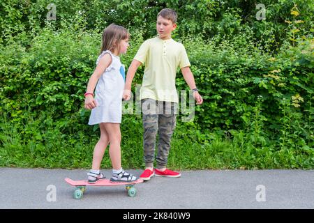 Une petite fille avec son frère se balade sur une planche à roulettes dans le parc. Un mode de vie sain Banque D'Images