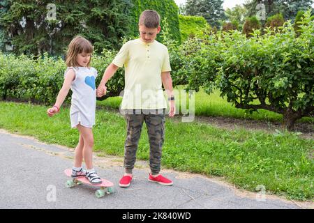 Grand frère enseigne le skateboard à petite sœur dans le parc, en tenant sa main Banque D'Images