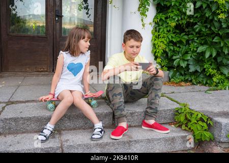 Les enfants jouent à l'extérieur. Un garçon et une fille sont assis sur les marches avec un téléphone dans leurs mains. Vêtements taille basse Banque D'Images