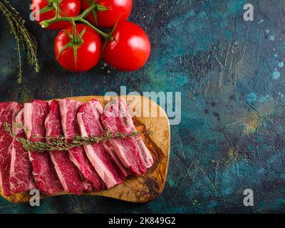 Sur fond de marbre foncé, une planche à découper en bois avec des morceaux de viande crus et une branche de romarin parfumé, tomates mûres. Cuisson des steaks de viande, repos Banque D'Images