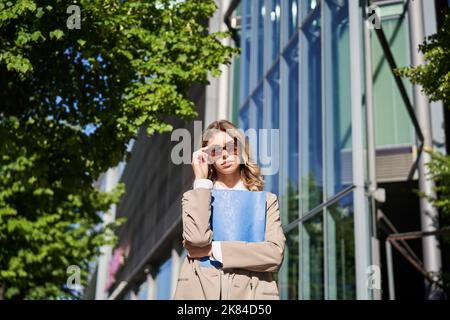 Portrait d'une femme de bureau confiante, femme d'affaires en lunettes de soleil posant à l'extérieur, près de son immeuble de bureau d'entreprise, portant un costume beige Banque D'Images