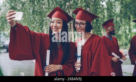 Les jolies filles qui terminent leurs études prennent le selfie avec des parchemins de diplôme à l'aide d'un smartphone, les jeunes femmes posent et sourient, leurs camarades célèbrent en arrière-plan. Banque D'Images