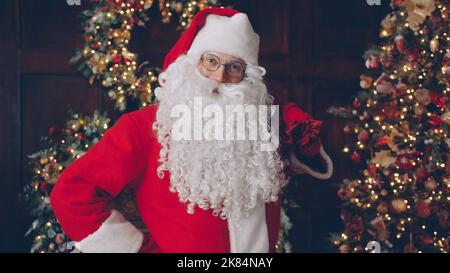 Portrait du joyeux Santa Clause sac de maintien avec des cadeaux près de l'arbre de Noël souriant et regardant l'appareil photo dans la maison décorée. Cadeaux et vacances concept. Banque D'Images