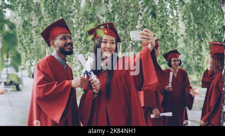 Les autres élèves prennent le selfie avec les diplômes posant et souriant, la fille tient le smartphone, les jeunes portent des robes et des chapeaux. Concept d'éducation et de style de vie moderne. Banque D'Images