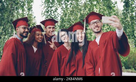 Des amis joyeux les étudiants diplômés prennent le selfie avec leur smartphone, les jeunes posent, montrent des gestes de main, font des visages amusants, sourient et rient. Banque D'Images