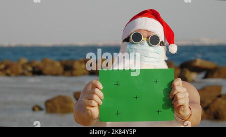 drôle de père noël, dans un masque de protection. Santa, dans des lunettes de soleil, portant un panneau vert, sur fond de mer. vacances du père noël pendant une pandémie du coronavirus ou un confinement, au bord de la mer. Photo de haute qualité Banque D'Images
