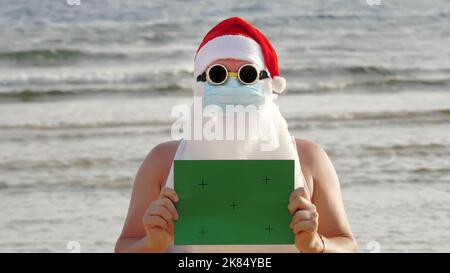 drôle de père noël, dans un masque de protection. Santa, dans des lunettes de soleil, portant un panneau vert, sur fond de mer. vacances du père noël pendant une pandémie du coronavirus ou un confinement, au bord de la mer. Photo de haute qualité Banque D'Images