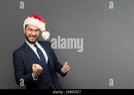 Portrait d'un homme d'affaires heureux et excité en chapeau de Noël riant contre fond gris de mur de studio. Concept de gain, de plaisir et de réussite Banque D'Images