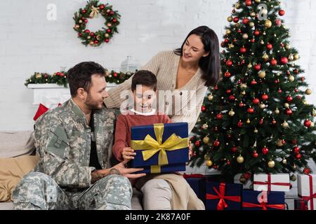 boîte cadeau d'ouverture d'enfant stupéfait près de père en camouflage et mère souriante dans le salon avec arbre de noël décoré Banque D'Images