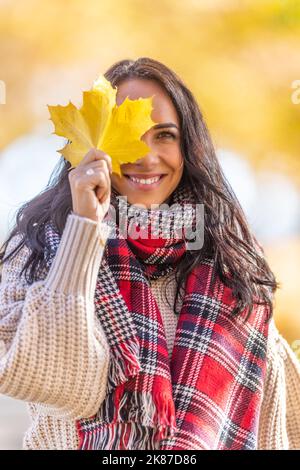 Une belle femme souriante vêtue d'automne tient une feuille jaune tombée à côté de son visage. Banque D'Images