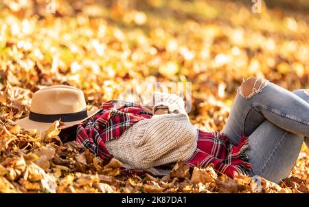 Jeune femme dormant ou se détendant dans le parc sur des feuilles mortes, tête couverte de chapeau pendant le chaud coucher de soleil d'automne. Banque D'Images