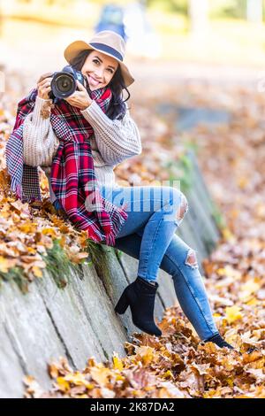 Une photographe féminine souriante et élégante, sous un chapeau et des vêtements d'automne élégants, prend des photos à l'extérieur lors d'une journée d'automne ensoleillée. Banque D'Images