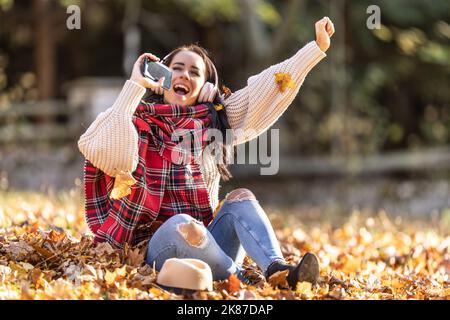 Une brunette amusante dans des vêtements d'automne élégants tenant le téléphone comme un micro chantant en automne laisse écouter de la musique avec des écouteurs. Banque D'Images