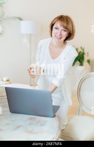 Jolie femme âgée debout près de son bureau avec un verre d'eau qui regarde l'appareil-photo sourire. Femme blanche adulte confiante travaillant à domicile Banque D'Images