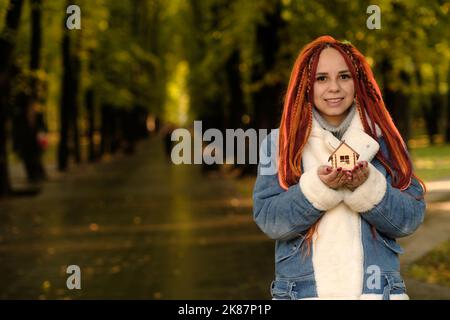 Bonne femme tenant le modèle à la maison dans le parc. Jeune femme positive avec des dreadlocks tenant une petite maison en bois souriant et regardant la caméra dans le parc d'automne. Banque D'Images