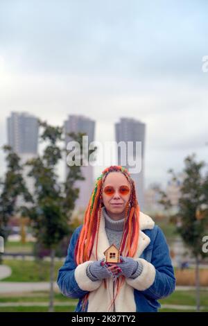 Jeune femme positive dans des vêtements chauds et avec de longues dreadlocks teints tenant une petite maison en bois et regardant la caméra contre des gratte-ciel flous dans ci Banque D'Images