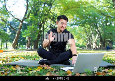 L'instructeur de fitness travaille en ligne utilise un ordinateur portable dans le parc assis en position lotus, l'athlète asiatique communique avec les étudiants à distance, la formation par vidéoconférence en ligne à distance Banque D'Images