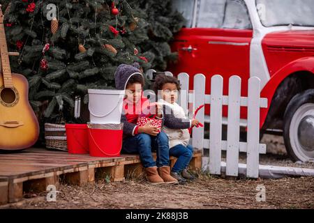 Les enfants afro-américains en vêtements d'hiver s'assoient sur des palettes en bois près de l'arbre de Noël, ouvrant des cadeaux. Garçon tient une boîte rouge dans les mains, une fille joue avec l'esprit Banque D'Images