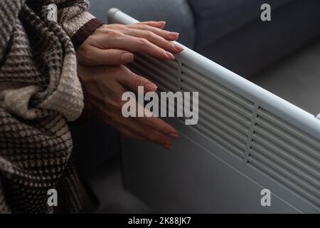 Une fille chauffe les mains gelées au-dessus du radiateur chaud, en gros plan. Femme portant un chandail en laine se réchauffant tout en étant assise près d'un radiateur de chauffage. Femme Banque D'Images