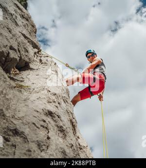 Grimpeur actif homme d'âge moyen dans un casque de protection regardant la caméra tout en descendant du mur de roche de falaise à l'aide d'une corde avec dispositif de belay et d'escalade ha Banque D'Images