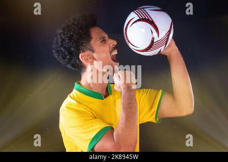 Joueur noir de football brésilien tenant le ballon et célébrant, isolé sur fond noir. Banque D'Images