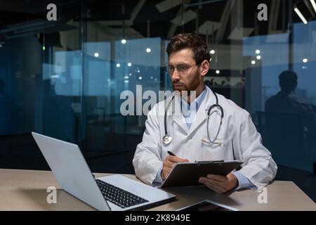 Médecin mature travaillant sur des travaux papier à l'intérieur d'un immeuble de bureaux miteux, homme en blouse médicale utilisant un ordinateur portable remplissant des documents médicaux en se concentrant et en pensant Banque D'Images