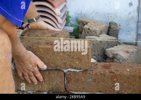 Images des mains d'un vieux maçon alors qu'avec du ciment, il construit un mur bas avec des briques de tuf. Homme au travail Banque D'Images