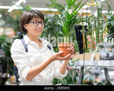 La femme choisit des plantes pour la maison. Étagères avec des semis, plantes à fleurs et graines dans la boutique de fleurs. Marché agronomique intérieur. Banque D'Images