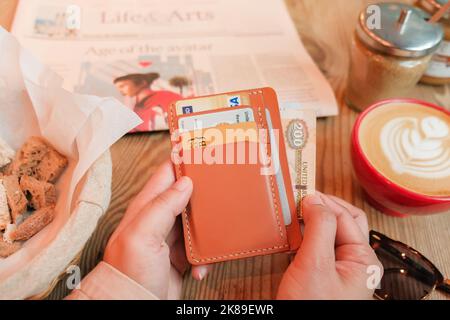 Main sort 200 eau Dirham de cuir Havane porte-carte avec pièces d'identité, cartes de débit, cartes de crédit. Café, panier à pain, journal, lunettes de soleil sur table en bois. Banque D'Images