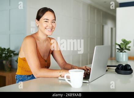 Je suis plus productive lorsque je travaille à la maison. Une jeune femme attrayante assise seule au comptoir de sa cuisine et utilisant son ordinateur portable. Banque D'Images