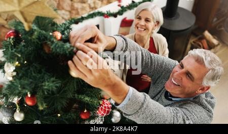 Ce n'est pas sur l'arbre, c'est autour de lui. Un heureux couple senior décorant leur arbre de Noël à la maison. Banque D'Images