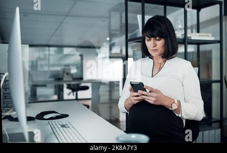 Rester à jour avec mon emploi du temps. Une femme d'affaires enceinte attrayante assise seule au bureau et utilisant son téléphone portable. Banque D'Images