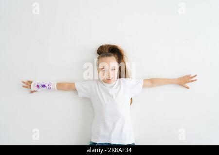 Vue de dessus de gaie petite fille mignonne avec cassé à la main enveloppé dans un bandage en plâtre avec dessin coloré couché sur le sol blanc en studio regardant l'appareil photo. Banque D'Images