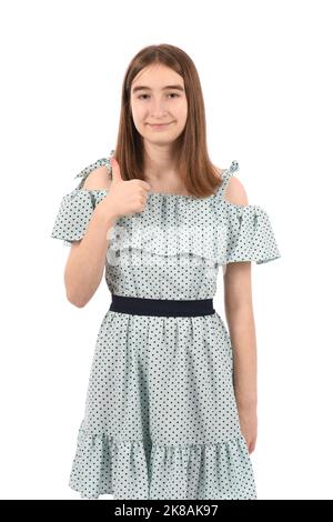 Jeune fille belle dans une robe bleue avec des points de polka sur un fond blanc, souriant à l'appareil photo et pouces vers le haut. Banque D'Images